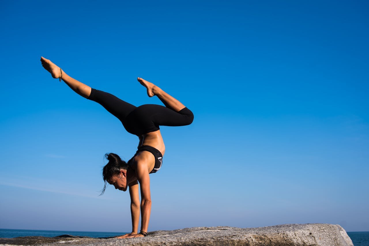 who-we-are A woman performing a yoga handstand on a rock by the sea under a clear blue sky.
